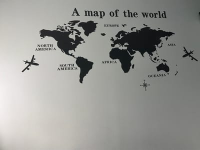 The image shows a wall decal of a world map on a light gray textured wall. The map is black and depicts the continents of North America, South America, Europe, Africa, Asia, and Oceania. Several airplane decals are also present, flying over the map. Text labels for the continents are in black sans-serif font. A compass rose graphic is located in the lower right quadrant of the map. The wall appears to be indoors, possibly in a home or office setting. There is no information in the image to suggest a location in Augsburg, Germany.