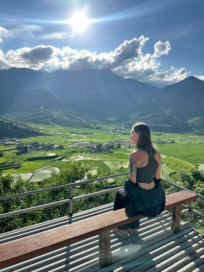 A person is seated on a wooden bench at an elevated viewpoint, overlooking the terraced rice paddies of Tú Lệ, Vietnam. The female subject, positioned with her back mostly to the camera, is looking leftward into the expansive green valley. She wears a dark olive crop top, dark pants, and a dark jacket draped over her lap, revealing a multi-colored, intricate tattoo on her left shoulder and upper arm. The bench rests on a corrugated metal platform, with a two-bar metal railing and green foliage partially obscuring the immediate foreground. Beyond, a vast landscape of vibrant green terraced rice fields with reflective water sections unfolds, interspersed with clusters of village buildings. Distant mountains rise from the valley, layered in a blue haze. The sky is bright blue with scattered white clouds, and a brilliant sun in the upper right casts a strong lens flare and sunbeams across the scene.