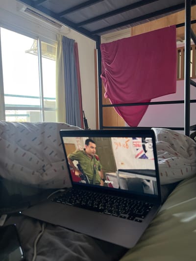 The image shows a laptop computer on a bed in a room located in the city of Beersheba, Israel (latitude: 31.249, longitude: 34.782).  The laptop is open and displaying a scene from a movie or television show. The scene depicts a man wearing a brown uniform, holding a rifle, standing in what appears to be a room. There are white walls in the background. The laptop is positioned on a bed with a white and light-brown patterned comforter.  A pink sheet or blanket hangs from the top bunk of a bunk bed. The bed is against a wall with a window and curtains. The window shows a view of the outside.  The room appears to be a bedroom.