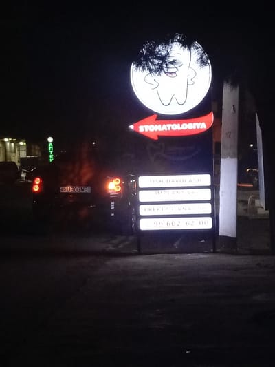 The media file captures a nighttime scene in what appears to be an urban or semi-urban area of Avval', Uzbekistan. The primary focus is an illuminated sign for a dental clinic, or "Stomatologiya" as indicated by the red arrow-shaped part of the sign.
The sign is composed of two main sections:
1. **Top circular section:** Features a cartoon-like illustration of a tooth with distinctive dark, curly hair or a wig, and a friendly expression.
2. **Red arrow section:** Below the circular emblem, a red arrow points to the left, bearing the white text "STOMATOLOGIYA", which translates to "Dentistry" or "Stomatology".
3. **Rectangular information panel:** Below the arrow, a vertical stack of four illuminated white rectangles displays services and contact information in Uzbek:
* "TISH DAVOLASH" (Tooth Treatment)
* "IMPLANT VA" (Implant and...) - likely referring to implant procedures.
* "BREKET O'RNATISH" (Braces Placement)
* "99 602 62 00" (A telephone number)
To the left of the sign, a dark-colored sedan, possibly a Chevrolet, is visible with its tail lights illuminated, suggesting it is either stopped, parked, or moving slowly. Its rear license plate clearly displays "40U300NB". In the blurred background, a building features a green neon sign that partially reads "ANTEY". Adjacent to the dental clinic sign on the right, a concrete post, possibly part of a fence or structure, is visible. The scene is illuminated only by artificial lights, consistent with the nighttime setting.