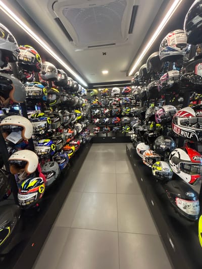 The image depicts the interior of a retail store specializing in motorcycle helmets, located in Alor Setar, Malaysia. The store is filled with numerous helmets displayed on shelves and racks, creating a dense and organized presentation. Helmets of various colors, designs, and brands are visible, ranging from full-face to open-face styles. The lighting is bright, illuminating the products and the store environment. The floor is tiled with a light gray pattern. The overall impression is one of a well-stocked and professionally managed establishment catering to motorcycle enthusiasts.