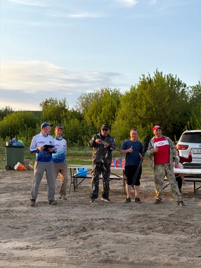 The image shows a group of six people standing outdoors, likely in the city of Krasnyi Bor, Russia, given that one of the team jerseys has the Russian flag on it. The ground appears to be sandy or dirt-covered. The weather is clear. In the background, there are trees and the rear of a white vehicle, where there's a person seated.
Five of the people are standing. Two men wear matching team shirts with the Russian flag on the sleeves. One man is dressed in camouflage clothing and is holding a small object, while another wears a blue shirt and stands with a black object, possibly a firearm. The last standing man wears camouflage pants and a red shirt with a white diamond.
At the rear of the vehicle is a person seated.