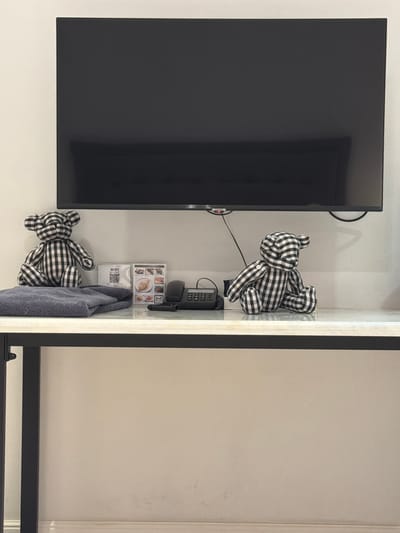 A black and white checkered teddy bear sits on a stack of folded gray towels on a marble-topped table. To the right of the towels, a small framed sign with a QR code and "WIFI" printed on it stands next to a framed picture menu. Further to the right, a black telephone with a coiled cord is positioned next to another checkered teddy bear, this one also sitting. Above the table, a large black flat-screen television is mounted on a white wall. The scene appears to be set in a hotel room in Ban Phonsavang Neua, Laos.