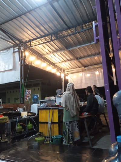 The image depicts an outdoor eatery, likely a warung or small restaurant, in Cangkring, Indonesia. The structure has a corrugated metal roof supported by a metal frame. String lights hang below the roof. In the background, there's a building with several windows.
The foreground shows the food preparation area on the left. Several gas canisters are visible, along with cooking equipment and supplies.
Several people are present: a woman with a head covering is standing near the preparation area, and a group of three or four people are seated at a table, seemingly eating or socializing. The seating appears to be basic, consisting of simple wooden stools.