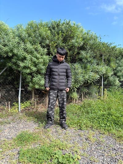 The scene is set outdoors in Icod de los Vinos, Spain, under a clear blue sky. A young male individual, appearing to be of East Asian descent, stands centrally in the frame, facing forward with his head tilted downward, gazing at the ground. He is dressed in a black, hooded puffer jacket, zipped up, and dark-colored pants with a repeated pattern of light-colored graphics. He wears dark athletic shoes and has white wireless earbuds visible in both ears. The ground directly beneath and around him is composed of dark gray and black gravel interspersed with patches of green, low-lying vegetation, which becomes denser towards the right side of the image. Behind the subject, a metal chain-link fence is partially visible, running horizontally and acting as a boundary for a dense, tall hedge of green, succulent-leafed foliage that forms the primary background. The sky above is predominantly clear and bright blue, with a few faint white clouds visible in the upper right quadrant. The scene is illuminated by natural, direct sunlight.