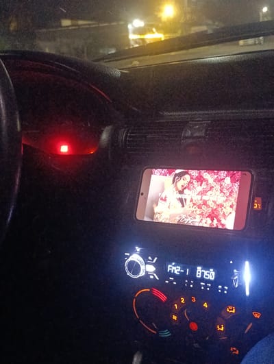 The image shows a close-up view of a car's dashboard at night. A smartphone is mounted on the dashboard, displaying a video of a woman with a floral background. The car's radio is visible, displaying "FM2-1 87.50". There are several illuminated buttons and dials on the dashboard, including what appears to be a temperature display showing "25 C". The overall lighting suggests the image was taken inside a moving vehicle. The exterior environment is dark, with some faint lights visible through the windshield. No specific location in Levakant, Tajikistan can be determined from the visual information.