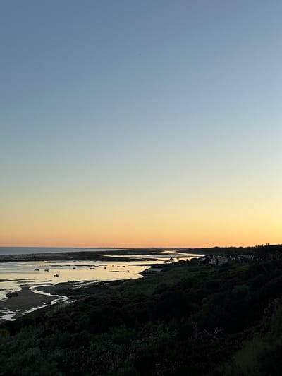 The image shows a landscape at what appears to be dusk or dawn. The sky transitions from a light blue on the left to an orange hue near the center. A body of water runs along the right side of the image, reflecting the light of the sky. The shoreline is bordered by dense vegetation. The location is Vila Real de Santo António, Portugal. Visible are sandy areas and water channels formed by the tide. The scene suggests a coastal environment. No people are visible in the frame.