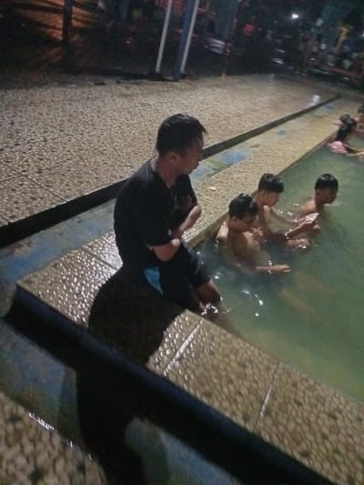The image captures a nighttime scene at what appears to be a natural hot spring or swimming pool in Kecamatan Pangalengan, Indonesia. In the immediate foreground, a man wearing a black t-shirt and dark shorts with a blue accent is seated on the tiled edge of the pool, his arms crossed. He is looking towards the water where three young boys are immersed up to their chests, seemingly relaxing or bathing. One boy, closest to the camera, looks downwards, while another glances towards the man.
Further into the pool, additional figures are faintly visible in the dim lighting, suggesting more people are enjoying the water. The area surrounding the pool is paved with wet, light-colored, cobblestone-like tiles. The overall illumination is low, indicating it is evening or night, with some artificial light sources casting soft glows in the background. A dark railing or barrier partially obstructs the lower left foreground. The atmosphere appears calm and leisurely, with individuals enjoying the warm water. No text is visible in the image.