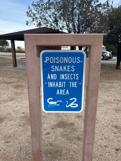 The image depicts a warning sign in the city of Dragoon, United States, indicating the presence of poisonous snakes and insects. The sign, which is blue with white lettering, is mounted on a brown metal post. The text on the sign reads "POISONOUS SNAKES AND INSECTS INHABIT THE AREA." Below the text, there are illustrations of a scorpion and a coiled snake. The background shows a dry, gravelly ground, a covered picnic structure with a brown roof, some trees, and a truck in the distance under a cloudy sky. The overall environment suggests an outdoor, possibly arid, location.