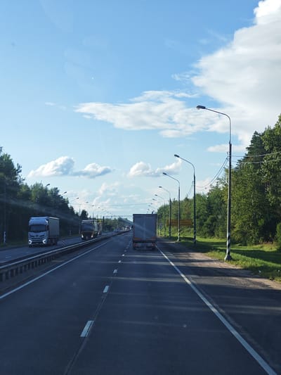 The image depicts a view from a moving vehicle on a multi-lane highway, likely in Staroye Melkovo, Russia, given the context. The sky is bright blue with scattered white clouds. On the right side of the road, a dense line of green trees borders the highway. Along the road, there are numerous streetlights on poles, some with wires connecting them. In the foreground, the asphalt road is visible with white lane markings. Several trucks are on the highway: a white truck is in the left lane, a yellow and white truck is in the middle lane, and a large gray trailer truck is in the right lane, partially visible. There are also other vehicles in the distance. A yellow sign with black lettering is visible on the right side of the road, and a speed limit sign (90) is also present. The scene suggests a daytime drive on a well-maintained highway.