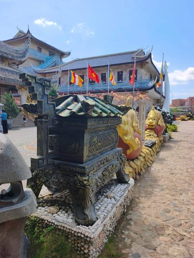 The image captures a scene at a Buddhist temple in Tuy An Nam, Vietnam, on a bright, sunny day with a partly cloudy sky. In the foreground, a large, dark, ornate incense burner with a green, tiled roof sits on a raised platform adorned with white pebbles and shells. To the right of the incense burner, a golden dragon statue is perched on a wall decorated with gold coins and more dragon figures. Behind this, a tall white statue of a Buddhist figure stands near a building with a dark tiled roof and traditional Vietnamese architectural elements, including curved eaves and ornate decorations. Several national flags, including the Vietnamese flag and the Buddhist flag, are displayed on poles in front of the temple buildings. The ground is paved with cobblestones. In the background, glimpses of other temple structures and some red-tiled roofs of nearby buildings are visible. A few people are present; one person in a blue shirt and black pants stands to the left, observing the scene, and a child in a dark dress is seen on a staircase in the background. Green grass and bushes are present around the base of the incense burner and to the left of the paved walkway.