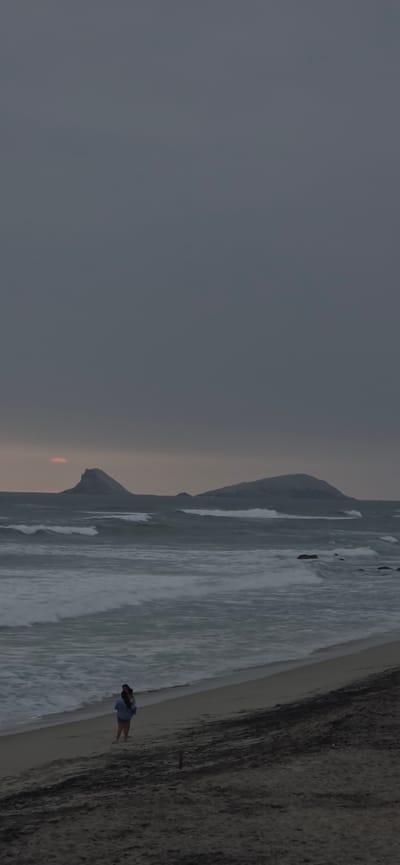 A person is standing on a sandy beach in Punta Hermosa, Peru. The person is wearing a blue top and shorts, and appears to be facing away from the camera. The ocean is in the background, with waves crashing on the shore. There are two rock formations in the distance, and the sky is overcast. The lighting suggests it is either dawn or dusk. The sand on the beach is a light brown color, and there are some dark patches of wet sand near the water. The overall mood of the image is serene and somewhat moody due to the overcast sky.