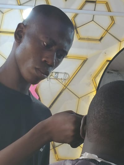 The image shows two people, seemingly a man and a child, in what appears to be an indoor setting with a decorative ceiling. The man, in the foreground and angled towards the camera, has dark skin and a shaved head. He is wearing a black t-shirt. His gaze is directed upwards and to the side. A portion of another person's head, also with dark skin, is visible in the upper right corner of the frame. The background features a ceiling with a geometric pattern of hexagonal shapes outlined in gold, illuminated by what seems to be artificial lighting. A metallic object, possibly a light fixture or a hook, is visible hanging from the ceiling. The scene is captured from a low angle, looking upwards. The lighting suggests it could be daytime or evening indoors. There is no visible text in the image. The overall impression is that of a candid or informal shot, possibly at a venue or home with distinctive architecture.