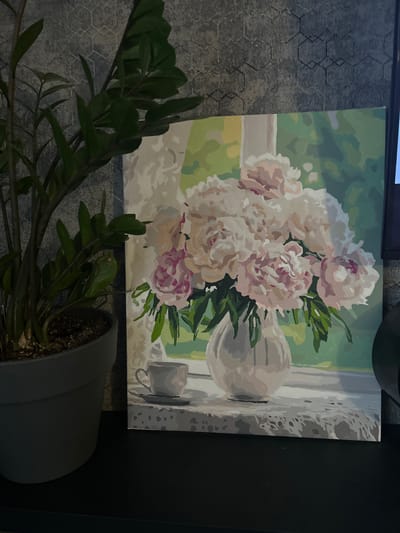 A still life painting of peonies in a vase is displayed on a black surface in front of a grey textured wall. To the left of the painting, a potted green plant with dark leaves is visible, its pot a muted grey. The painting itself depicts a bouquet of light pink and white peonies in a rounded white vase. A white teacup and saucer are placed to the left of the vase on a surface covered with a white lace doily. Behind the vase and flowers, sheer white curtains are suggested, with a soft green and yellow light filtering through. The background of the painting suggests a window with a garden view. The overall scene is dimly lit, with the painting being the primary illuminated subject. No people are present in the image.