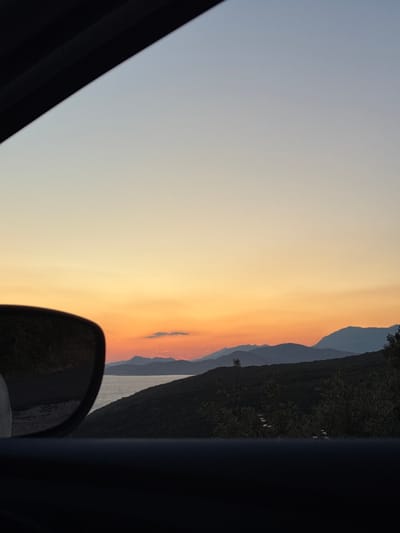 The image captures a scenic view through a car window at sunset. The sky transitions from a pale blue at the top to a warm orange and yellow gradient near the horizon. Silhouetted mountains form a jagged line against the colorful sky. In the foreground, a car's side mirror reflects a dark road and vegetation. Below the horizon, lush green hills and a body of water, likely the Adriatic Sea, are visible. The location is specified as Glavatičići, Montenegro.