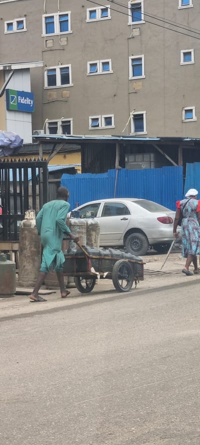 In Alagbede, Nigeria, a man in a teal robe is pushing a cart laden with dark-colored containers along a paved road. He is walking away from the viewer, his bare feet clad in sandals. Beside him, a woman in a patterned dress and white headscarf walks in the same direction, leaning on a thin pole. Behind them, a silver sedan is parked on the road. In the background, a building with a gray facade features numerous windows, some open and some closed, and a blue sign with the "Fidelity" logo. The street scene is bordered by a fence and some structures with corrugated metal roofing.
