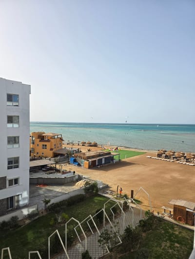 A high-angle, landscape-oriented photograph depicts a view of a beach and coastline in Al Ahia', Egypt. The image is composed of a clear, light blue sky in the upper left quadrant. Adjacent to the sky is the light blue/green ocean, marked with several people kite surfing in the distance. The beach is sandy, and on its right side is a row of beach umbrellas and beach chairs.

Between the beach and the foreground are several buildings. The buildings range in height and color, from tan to yellow. Adjacent to the buildings is a verdant, green area of grass, with a person walking along the edge of the beach. Near the bottom of the picture is a white, multi-story building. The photograph is taken in daytime. The image appears slightly rotated counterclockwise.