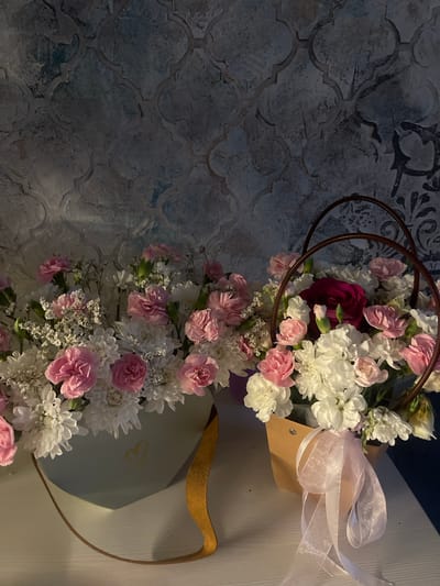Two floral arrangements are displayed on a white surface against a textured, dark grey and blue patterned wallpaper. The arrangement on the left is in a light blue, heart-shaped box with a gold heart emblem and a tan strap. It contains an abundance of pink and white carnations, along with small white flowers. The arrangement on the right is in a brown paper bag and features a dark pink rose as a focal point, surrounded by pink and white carnations and other white flowers. This arrangement has a circular, dark brown wire handle. A sheer pink ribbon is tied around the front of the brown paper bag. The lighting suggests an indoor setting, likely a room in Noyabrsk, Russia. No people are present in the image.