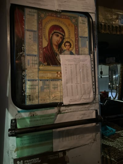 The image displays a dimly lit interior scene, likely within a public transport vehicle such as a bus or minibus, situated in Kapitanivka, Ukraine. The central subject is a vibrant religious icon of the Virgin Mary holding the Christ Child, which also functions as a calendar, with "202" visible, indicating a year such as 2022 or 2023. This icon is placed behind a clear plastic sleeve.

Various documents and schedules are affixed around the icon, some also protected by plastic sleeves. A vertical metal pole or bar is visible on the right side of the frame. The setting appears to be a functional space for passenger transport, personalized with a religious image.

Notable visible text, primarily in Ukrainian, includes "Шановні пасажири" (Dear passengers), indicating a public service. Further text specifies the service provider: "Вас обслуговує ТОВ «Центр" (You are served by LLC "Center"), along with an address: "с. Горенен, вул. Соборна 20" (village Horenen, Soborna street 20). A partial logo or banner on the far right reads "ГВАРДІЯ НАСТУПУ" (Guard of the Offensive/Attack). The dim lighting suggests the image was taken at night or in an enclosed space with limited natural light.