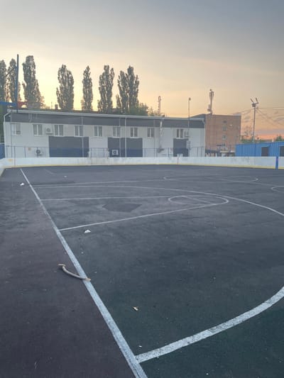 The image depicts an outdoor basketball court with dark asphalt surfacing and white boundary lines. In the background, a multi-story building with a white and grey facade stands adjacent to a row of tall, leafy trees. The building has numerous windows and visible air conditioning units. The scene is bathed in the warm light of either sunrise or sunset, casting a soft glow and creating long shadows. The sky is a gradient of pale orange and grey, suggesting a calm and possibly cool atmospheric condition. On the court, there are some scattered pieces of litter. A thin, curved twig or piece of debris lies on the court near a white line. The overall impression is one of a quiet, urban recreational space during a transitional time of day. There is no visible text in the image.