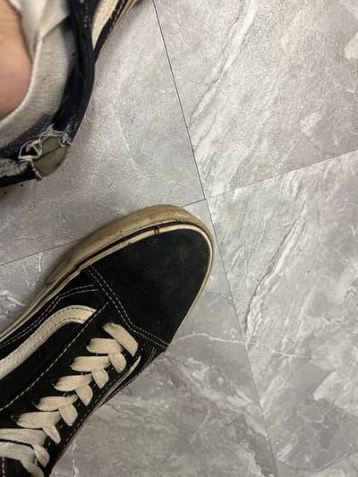 The image shows a close-up, overhead view of two worn, black Vans sneakers on a gray, marbled tile floor. One shoe, on the left, is partially visible and appears to have a torn section near the ankle opening, revealing white fabric underneath. The other shoe, more prominently featured on the right, shows the toe box and laces of a black canvas Vans sneaker with a white side stripe and white laces. The white rubber sole of this shoe is dirty and shows signs of wear. A thin, dark line, likely a grout line between tiles, runs diagonally across the image. There are no people visible, and the scene appears to be indoors. The image provides no information about the location in the city of Ленинский, Russia.