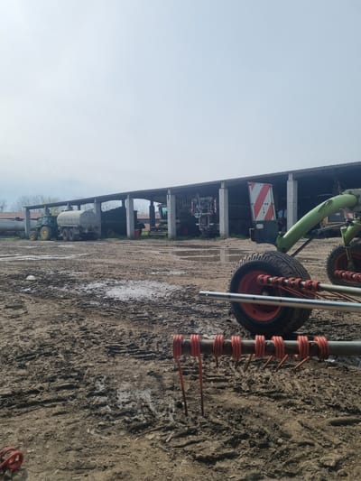 The image shows a Claas Volto tedder parked on a muddy dirt surface in Ovcharovo, Bulgaria. Behind it, there is a shed with farming machinery inside. A light-gray sky is visible above.