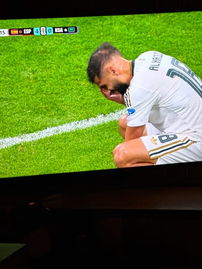A television screen displays a soccer match with a score of ESP 4 - 0 KSA. A player in a white jersey with black and gold trim is seen kneeling on the green grass, with his face buried in his hands. His jersey has the name "ALHARBI" and the number "10" on the back. The image is taken from a slightly elevated angle, with the lower portion of the screen showing the dark interior of a room, suggesting the TV is in a home setting. No specific location within Bykovo, Russia, is visible or discernible from the image.