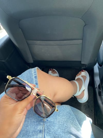 The image shows a close-up view of a person's legs and hands inside a vehicle, likely in the city of Tyulyugul, Russia. The person is wearing a denim skirt and white platform sandals, and holding a pair of sunglasses. The background consists of the dark fabric of a car seat. The lighting suggests it is daytime, possibly sunny. The overall scene is casual and personal, capturing a moment inside a car.