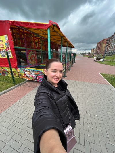 In Medvedevo, Russia, a woman is taking a selfie outdoors on a cloudy day. She is wearing a black puffer jacket and a small pink purse with silver studs, which is hanging from her shoulder. She is smiling at the camera. Behind her, to the left, is a colorful children's play structure with a red and yellow canopy. The play structure is enclosed by a green metal fence and is decorated with cartoon characters. A paved walkway with red and gray bricks extends from the foreground towards the background, lined with green grass. Further in the background, apartment buildings are visible on the right side of the frame, along with streetlights and park benches. The sky is overcast with heavy, gray clouds.