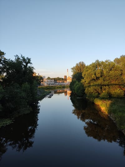 The image captures a serene river scene in Kolín, Czechia, bathed in the warm glow of the setting sun. The Elbe River dominates the foreground, its calm surface reflecting the clear blue sky and the lush greenery lining its banks.

On the left bank, dense trees cast long shadows, their branches reaching towards the water. On the opposite bank, a row of modern white buildings stretches along the river, their reflections shimmering on the water's surface. Further in the distance, an industrial complex with a tall, red-and-white striped chimney stands as a stark contrast to the natural beauty of the surroundings. A small wooden dock juts out into the river, hinting at recreational activities.

The overall impression is one of tranquility and urban integration, where nature and industry coexist under a vast, unblemished sky.
