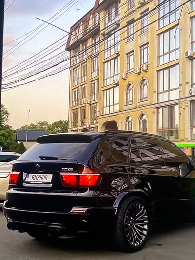 In Rokytne, Ukraine, a black BMW X5 M is parked on a street. The rear of the vehicle is visible, featuring its taillights and license plate, which reads "BMWHUB". The car is parked in front of a multi-story building with yellow facade and numerous windows. Overhead, a network of electrical wires crisscrosses the sky. The overall scene suggests an urban environment in daylight.