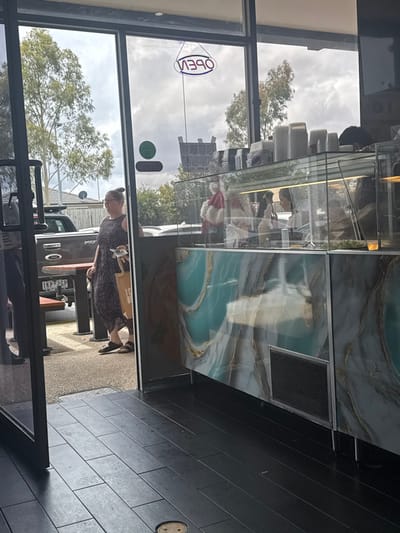 The image, taken from inside a commercial establishment and oriented sideways, looks out through a large window onto an outdoor scene. Inside, reflections on the window reveal a display counter with a distinct light blue, gold, and brown marble-like pattern, as well as a dark tiled floor and a visible vent underneath the counter.

Outside, a woman wearing a dark, possibly polka-dotted, dress with her hair in a bun walks past the window, carrying what appears to be an item in her hand. Behind her, a light-colored utility vehicle or truck is visible, displaying a license plate that reads "1KP 2GP". The background features trees, an overcast sky, and hints of distant structures. A blue, neon-style "OPEN" sign is reflected in reverse on the window, indicating it is mounted to be read from outside. The scene takes place during the daytime, under cloudy weather conditions. The overall setting suggests a commercial street or area in Carrum Downs, Australia.