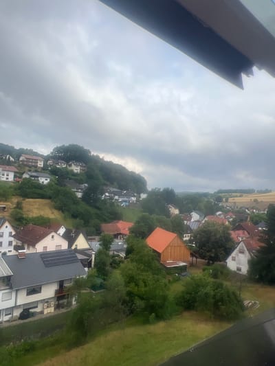 This image depicts a view of the village of Ober-Mumbach, Germany, on an overcast day. The scene is dominated by a hillside covered with trees and dotted with houses. In the foreground, several houses are visible with red and gray tiled roofs. One house in the middle left has solar panels on its roof. Further up the hill, more houses with various architectural styles and roof colors are scattered amongst the greenery. The background shows a wider landscape with more trees and fields, suggesting a rural or semi-rural setting. The sky is heavily clouded, casting a diffused light over the entire scene. No people are visible in the image.