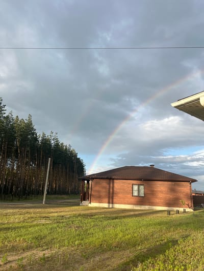 The image captures a rural scene in Mokhovatka, Russia, dominated by a double rainbow arching across a cloudy sky. A modern, red-brick house with a dark brown roof is prominently featured in the foreground, positioned to the right of the frame. A single window is visible on the side of the house. To the left, a dense forest of tall, slender pine trees lines the horizon. A rustic wooden utility pole stands in the grassy field between the house and the forest. The field itself is covered in green grass, with visible patches of bare earth and a tire track, suggesting recent activity. The lighting indicates it is likely late afternoon, with sunlight casting long shadows.