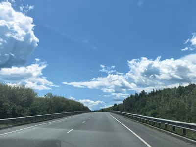 The image shows a highway in Vozrozhdenie, Russia, under a bright blue sky with scattered clouds. The road is paved and has white lane markings. On either side of the road, there are guardrails, and beyond them, lush green trees form dense forests. The trees appear to be deciduous, with full canopies of leaves. The sky is a vibrant blue, with fluffy white cumulus clouds drifting across it. The overall impression is one of a scenic drive through a natural landscape.
