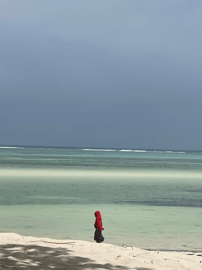The image depicts a beach scene in Kiwengwa, Tanzania. A single individual stands on white sand at the water's edge, facing right. This person wears a vibrant red head covering and a dark, full-length garment. The calm, shallow ocean water, exhibiting shades of turquoise and light green, extends into the midground, with faint whitecaps visible further out, indicating distant waves or a reef. The sky above is a dark, overcast grey-blue. The foreground shows shaded areas of sand with minimal debris, likely from unseen overhead vegetation. No discernible actions or interactions beyond standing are observed.