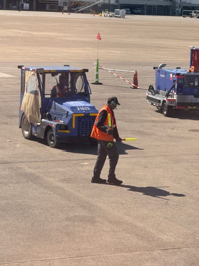 The scene depicts active ground operations on an airport tarmac at Nashville International Airport (BNA) in Nashville, Tennessee, United States of America, under bright, sunny conditions.
In the foreground, a male airport ground crew member, appearing to be of African descent, walks from left to right. He is dressed in a black baseball cap, black headphones with a microphone, an orange high-visibility safety vest over a dark long-sleeved shirt, dark pants, and dark shoes. He holds two fluorescent yellow/green marshalling wands in his hands. A distinct, elongated shadow of the man is cast on the light grey concrete pavement to his right.
To the left of the walking crew member, a blue airport tug vehicle, identified by the numbers "21025" and the "TUG" logo on its front, is stationary. Its cab is partially enclosed with clear windows and a tan canvas side covering. Inside the cab, a second male, also appearing to be of African descent and wearing an orange high-visibility vest, is seated as the driver.
In the mid-ground, to the right of the walking crew member, a blue wheeled utility unit, branded with "Southwest" and "TUG," is stationary. This unit features a red cover on its top surface. A safety barrier, consisting of a red and white striped pole, is suspended between a green and white striped pole and a red traffic cone. A small red flag is affixed to the top of the green and white striped pole by a thin wire. The background shows distant airport infrastructure and additional vehicles.