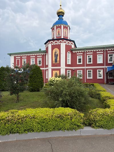 This is a medium shot, eye-level, exterior photograph of a red brick building with white trim, featuring a blue-domed tower topped with a golden cross. The building has multiple rectangular windows and ornate architectural details. On the tower, there are framed religious icons. In front of the building, there is a manicured garden with green bushes, a small tree with red berries, and flowering shrubs. The sky is overcast with clouds. The photograph appears to have been taken on a cloudy day. The building is identified as being located in the city of Norja, Russia.