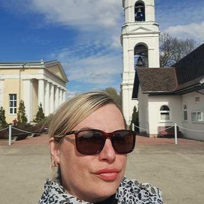 A selfie photograph shows a blonde woman wearing sunglasses. Behind her are two buildings against a blue sky with white clouds. The building on the left is a bell tower with a white facade and arched openings revealing bells. The building on the right has a light yellow facade, white columns, and a triangular pediment. The setting is Ivashevo, Russia.
