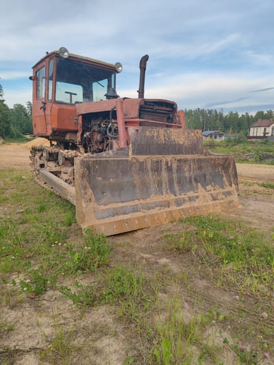 An orange tractor is parked in an outdoor area with a dusty ground and scattered grass. The tractor is a heavy-duty vehicle with a large metal blade at the front, covered in dirt. It has a driver's cab with windows and two headlights on top. The engine is partially exposed at the front of the tractor. In the background, there are trees, houses, and a cloudy sky. The location is described as the city of Zashchita, Russia. There are no people visible in the image.
