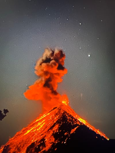 The image depicts a volcanic eruption. Orange lava flows down a slope, accompanied by a plume of smoke or ash. The background is a dark, speckled expanse, possibly the night sky with stars. No people or specific locations can be identified. The city of Placencia, Belize is not mentioned.