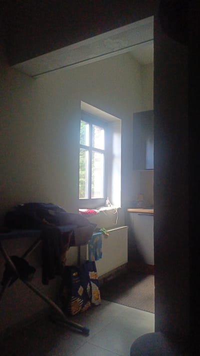 The image depicts a dimly lit interior scene. A prominent window with multiple panes is visible, allowing natural light to enter the room. Outside the window, greenery and the facade of a brick building can be seen. On the windowsill, a red cloth or rug is present, along with some wires. In the foreground, to the left, a clothes rack with dark clothing draped over it is situated. Next to the rack, a blue and yellow patterned bag or backpack rests on the floor. A white radiator is positioned below the window. To the right of the window, a section of kitchen cabinetry with a countertop is visible, suggesting a domestic setting. The overall environment appears to be a room in Nikolino, Russia.