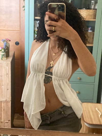 The image shows a person taking a mirror selfie. The person has curly dark hair and is wearing a white halter top with ruffles and a gold necklace with a cross pendant. They are also wearing a ring on their left hand. The background appears to be a room with light blue cabinets and wooden furniture. The lighting suggests it is daytime, possibly indoors. The overall mood seems casual and relaxed.