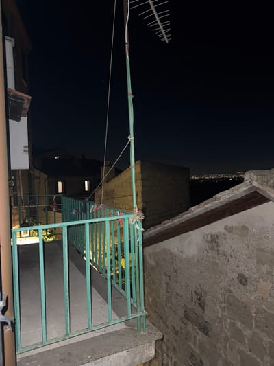 An outdoor scene taken at night in Castelnuovo di Porto, Italy. In the foreground is a metal railing painted light green. Clothes with clothespins are hung up to dry on the railing. A building overhangs the railing. In the background, buildings are dimly lit with windows visible. An antenna is seen against the night sky. Faint city lights are visible in the distance.