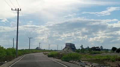 The image depicts a road curving through a semi-arid landscape under a cloudy sky. On the left, a tall utility pole with multiple crossbars supports overhead wires. The road, paved and marked with white lines, winds from the foreground into the distance. Lush green vegetation lines the left side of the road, with scattered bushes and trees further back. On the right side, the terrain is more barren with patches of dry grass and small rocks. In the mid-ground, a man-made structure, appearing to be a metal framework or partially constructed bridge, is situated atop a mound of earth. The background features a line of trees and utility poles against a sky filled with a mixture of white and gray clouds. The location is identified as Pusong, Indonesia. No people are visible in the frame, and no specific actions or interactions are captured. The overall scene is one of an outdoor environment, possibly a rural or developing area, with infrastructure elements and natural vegetation.