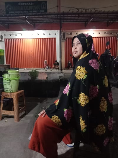 In Sekopek Wetan, Indonesia, at night, a woman wearing a black hijab with a floral pattern of pink and yellow flowers sits facing right. She is wearing an orange skirt. In the background, a row of orange roll-up doors is visible, suggesting a commercial or shop setting. To the left of the woman, there is a brown woven plastic stool with a green bucket on top. Further in the background, several other people are seated in front of the closed doors, some appearing to be engaged in activities. The overall lighting suggests it is evening or nighttime.