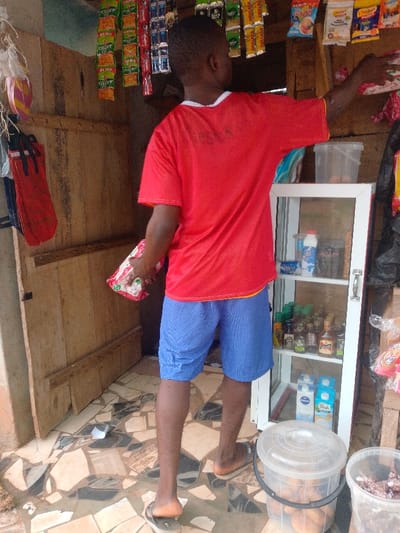 The image captures the interior of a small retail shop or provision stall in Umuoji, Nigeria. A male individual, viewed from the back, stands centrally within the frame. He is dressed in a red short-sleeved t-shirt, blue textured shorts, and brown flip-flops. His left hand holds a clear plastic package containing a white, granular product. His right arm is extended upward and to the right, in proximity to several hanging packaged goods.
The shop's walls on the left and background are constructed from vertical wooden planks. Numerous small, individually packaged items, including snacks and sachets (e.g., "Forever Crisp," "Milo," "Golden Penny," "Chikki Noodles"), are suspended from overhead lines or hooks across the upper wall sections.
To the right, a white display cabinet with glass doors contains multiple shelves stocked with various goods, including milk cartons (e.g., "Peak"), bottled items resembling sauces or spirits, and other packaged products. A clear plastic container filled with eggs rests on top of this cabinet. At floor level, two clear plastic buckets are visible; one contains spherical brown items, and the other, partially cropped, holds darker, irregular contents.
The floor is tiled with an irregular mosaic pattern of light brown, dark brown, and black pieces. Minor debris, including a small piece of white paper, is present on the floor. On the left wall, several red fabric items with black straps are hanging.