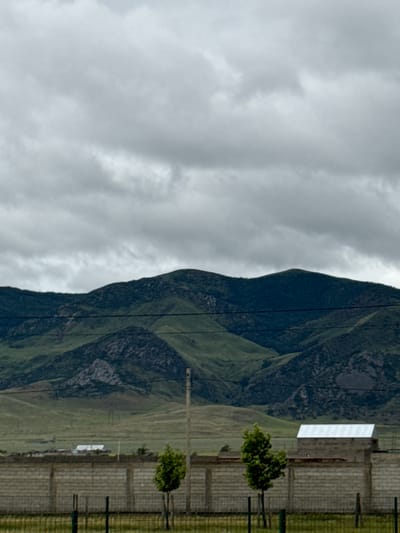 The image displays a mountainous landscape under an overcast sky, likely in Korkmaskala, Russia. The left side of the frame is dominated by a dense, grey, cloudy sky. On the right, large mountains or hills rise, their slopes covered in a patchwork of dark green foliage, lighter green grassy areas, and visible rocky sections.

In the mid-ground, towards the lower right, human infrastructure is visible. A prominent, long, rectangular building with a white or light-colored facade and a flat roof stands at the base of the mountains. In front of this building, a sturdy fence or wall constructed from light-colored bricks or concrete blocks extends horizontally. A wire or mesh fence runs parallel to this wall, closer to the viewer. Two small, deciduous trees with vibrant green leaves are positioned in front of the brick wall. A thin, vertical utility pole or mast stands behind the fences. Several thin, dark lines, possibly power lines or cables, traverse the scene, cutting across the sky and mountain slopes.

The time of day appears to be daytime, but the heavy cloud cover prevents direct sunlight and creates a muted, subdued lighting. There are no people visible, and no specific activity or event is taking place, presenting a tranquil yet somewhat somber scene. No visible text is present in the image.
