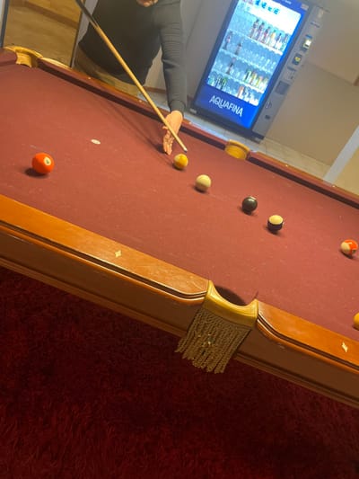 A billiard table with a red surface dominates the frame. Several billiard balls are scattered across the table: a striped 15 ball near the top, a 1 ball, a 2 ball being aimed at, and others in a line. A person holds a cue stick, preparing to strike the 2 ball. An Aquafina vending machine is visible in the background on the left side. Part of a red carpet is visible on the right side of the image.