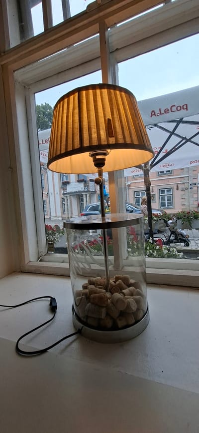 A table lamp with a pleated, tan lampshade is positioned on a white windowsill. The lamp base is a clear glass cylinder filled with wine corks. A black cord with an on/off switch is visible extending from the base. Through the window, a street scene in Kuressaare, Estonia is visible. The buildings are light-colored with classical architectural elements. There are umbrellas with a logo that appears to say "A.LeCoq". Cars and a person are visible on the street. Red and white flowers are planted in window boxes outside the buildings. The sky is overcast.